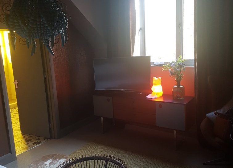 A cozy room with a TV on a cabinet beneath a window, a glowing bear-shaped lamp, a potted plant, a round wicker chair, two small stools, and a decorative chandelier. Light enters from the open door and window.