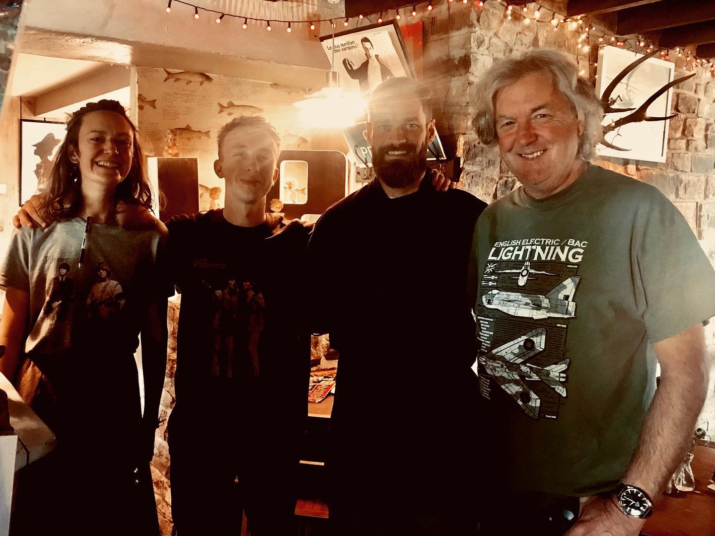 Four people stand closely together inside a warmly lit room, smiling at the camera. The background features string lights, framed pictures, rustic decor, and a menu reminiscent of something James May might appreciate.