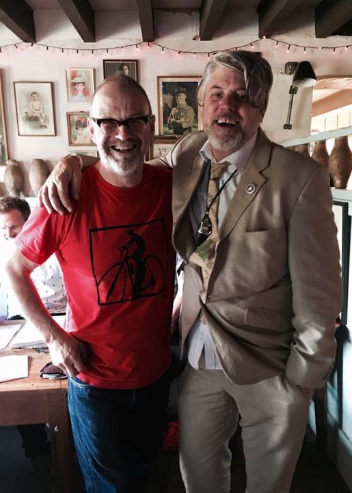 Two smiling men pose indoors; one wears a red graphic T-shirt and glasses, the other a beige suit with a pendant. Channeling Neighbours vibes, they stand arm in arm in a room decorated with pottery—one reminiscent of Joe Mangle.