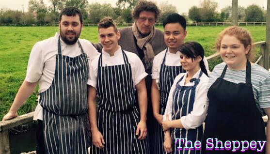 A group of six people, some in striped aprons and chef uniforms, stand together with Marco outdoors on a wooden bridge, green fields behind them. The words The Sheppey appear in purple text at the bottom right.