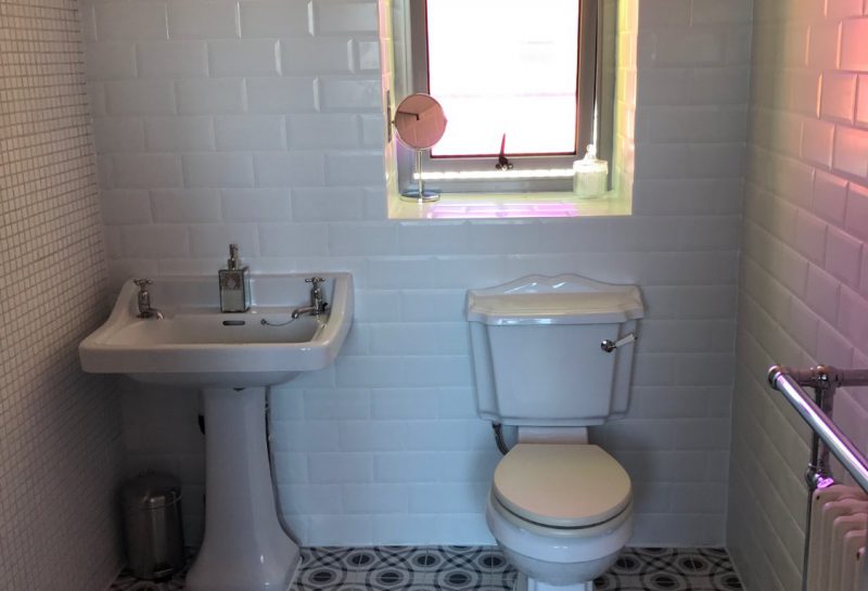 A small bathroom with white tiled walls, patterned floor tiles, a pedestal sink, toilet, wall-mounted towels, a radiator, and a window above the toilet letting in sunlight.