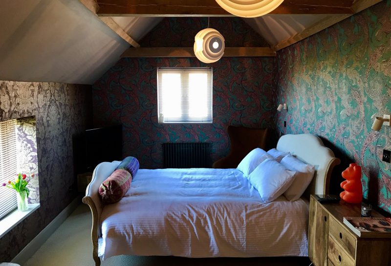 A cozy bedroom with patterned wallpaper, a white bed with pillows, bedside tables, two modern pendant lights, and a window with blinds letting in sunlight. Fresh flowers sit on the window ledge.