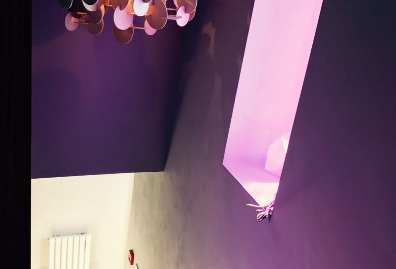 A modern staircase with a wooden handrail, patterned floor tiles, and a geometric chandelier hanging from the ceiling. Soft pink light glows from a vertical window near the stairs.