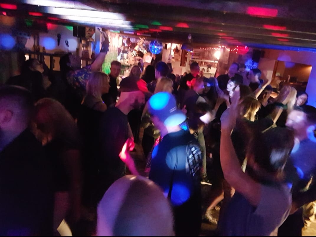 A crowded nightclub or bar, like the Sheppey Soul Shack, pulses with people dancing under colorful, dim lighting. Blue, red, and purple lights create a lively atmosphere as partygoers move and raise their arms.