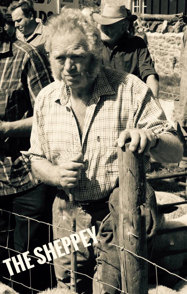 An older farmer with tousled hair and sideburns, wearing a plaid shirt, stands by a wooden post and wire fence at an outdoor event. The image is black and white. The text THE SHEPPEY appears in the lower left corner.