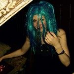 A person wearing a bright blue wig and a black sleeveless top sits indoors, partially obscured by a decorative frame. Smiling and holding part of the wig, they enjoy the dim lighting, capturing the festive spirit of New Years Eve 2019. A person wearing a bright blue wig and a black sleeveless top sits indoors, partially obscured by a decorative frame. Smiling and holding part of the wig, they enjoy the dim lighting, capturing the festive spirit of New Years Eve 2019.