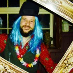 A smiling person with a blue wig, black top hat, floral lei, and red plaid shirt poses holding a large ornate picture frame indoors, celebrating New Years Eve 2019, with a window and festive decorations in the background. A smiling person with a blue wig, black top hat, floral lei, and red plaid shirt poses holding a large ornate picture frame indoors, celebrating New Years Eve 2019, with a window and festive decorations in the background.