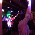 A band performs live on stage in a dimly lit venue with colorful lights shining. A guitarist wearing a hat stands in the foreground. Audience members, celebrating New Year's Eve 2019, are visible—one with an arm raised, enjoying the music. A band performs live on stage in a dimly lit venue with colorful lights shining. A guitarist wearing a hat stands in the foreground. Audience members, celebrating New Year's Eve 2019, are visible—one with an arm raised, enjoying the music.