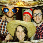 Four people pose together holding an ornate picture frame, dressed in playful New Years Eve 2019 costumes—oversized sunglasses, a straw hat, a red-and-white beanie, and round joke glasses—all smiling and enjoying themselves. Four people pose together holding an ornate picture frame, dressed in playful New Years Eve 2019 costumes—oversized sunglasses, a straw hat, a red-and-white beanie, and round joke glasses—all smiling and enjoying themselves.