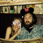 A woman holding stick-on shutter shades and a smiling man with a beard wearing Minnie Mouse ears pose together behind a large ornate picture frame, celebrating New Years Eve 2019 with various decorative items on a shelf behind them. A woman holding stick-on shutter shades and a smiling man with a beard wearing Minnie Mouse ears pose together behind a large ornate picture frame, celebrating New Years Eve 2019 with various decorative items on a shelf behind them.