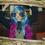 A person poses inside an ornate picture frame at a New Years Eve 2019 party, wearing a bright blue wig, oversized orange sunglasses, a pink feather boa, and a star necklace. The background shows a window and wooden paneling. A person poses inside an ornate picture frame at a New Years Eve 2019 party, wearing a bright blue wig, oversized orange sunglasses, a pink feather boa, and a star necklace. The background shows a window and wooden paneling.