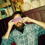 A bearded person in a plaid shirt wears purple shutter shades, holds a fake mustache and a lollipop, posing playfully in front of a shelf with vintage decorations and figurines—ready for a fun New Years Eve celebration. A bearded person in a plaid shirt wears purple shutter shades, holds a fake mustache and a lollipop, posing playfully in front of a shelf with vintage decorations and figurines—ready for a fun New Years Eve celebration.
