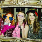 Three young women smile inside a large ornate picture frame, wearing colorful costume accessories like oversized glasses and feather boas, posing for a fun, festive New Years Eve 2019 photo. Three young women smile inside a large ornate picture frame, wearing colorful costume accessories like oversized glasses and feather boas, posing for a fun, festive New Years Eve 2019 photo.