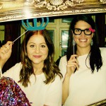Two women smile at the camera while holding a large ornate picture frame and playful props, capturing festive New Years Eve spirit. One wears a blue crown on a stick, the other novelty glasses with a pink bow—both dressed in white tops, celebrating 2019. Two women smile at the camera while holding a large ornate picture frame and playful props, capturing festive New Years Eve spirit. One wears a blue crown on a stick, the other novelty glasses with a pink bow—both dressed in white tops, celebrating 2019.