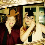 Two women pose playfully behind a large ornate picture frame, each holding a paper hat and a stick-on mustache to their faces. Smiling, they celebrate New Years Eve 2019 indoors in front of a window and decorated shelves. Two women pose playfully behind a large ornate picture frame, each holding a paper hat and a stick-on mustache to their faces. Smiling, they celebrate New Years Eve 2019 indoors in front of a window and decorated shelves.