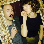 A man holding a fake mustache on a stick and a woman sit together inside an ornate, empty picture frame, playfully posing for the camera. The woman appears to be whispering to the man during a festive New Years Eve celebration. A man holding a fake mustache on a stick and a woman sit together inside an ornate, empty picture frame, playfully posing for the camera. The woman appears to be whispering to the man during a festive New Years Eve celebration.