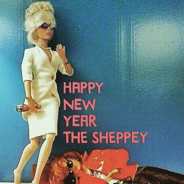 A doll in a white suit and sunglasses stands next to another doll lying on the floor in a red outfit. Pink text says HAPPY NEW YEAR THE SHEPPEY 2019 against a blue background, capturing a fun New Year's Eve vibe.