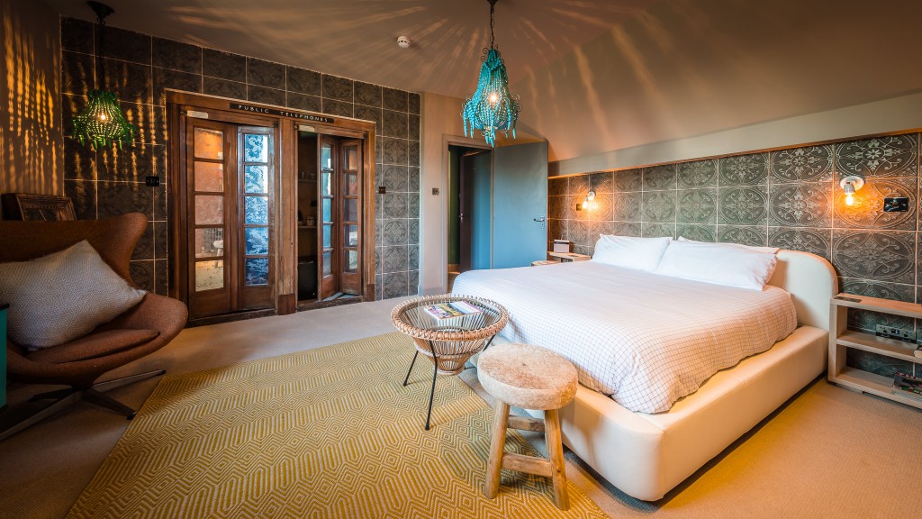 A cozy bedroom with a large white bed, patterned gray accent wall, warm lighting, a yellow rug, wicker furniture, and two blue hanging lamps. Wooden double doors and a brown armchair are also visible.