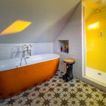 A bathroom with a sloped ceiling features an orange bathtub, patterned tile floor, a small black-and-wood stool, and a walk-in shower with yellow tiles and built-in shelves holding toiletries.