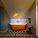 A bright bathroom with a freestanding orange bathtub beneath a sloped ceiling, patterned floor tiles, white subway-tiled walls, a shower to the right, and a bear-shaped side table holding a wooden tray.