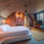 A cozy bedroom with a large white bed, bedside tables, patterned accent wall, chandelier, artwork above the bed, a TV on a console, large window with dark curtains, and warm ambient lighting.