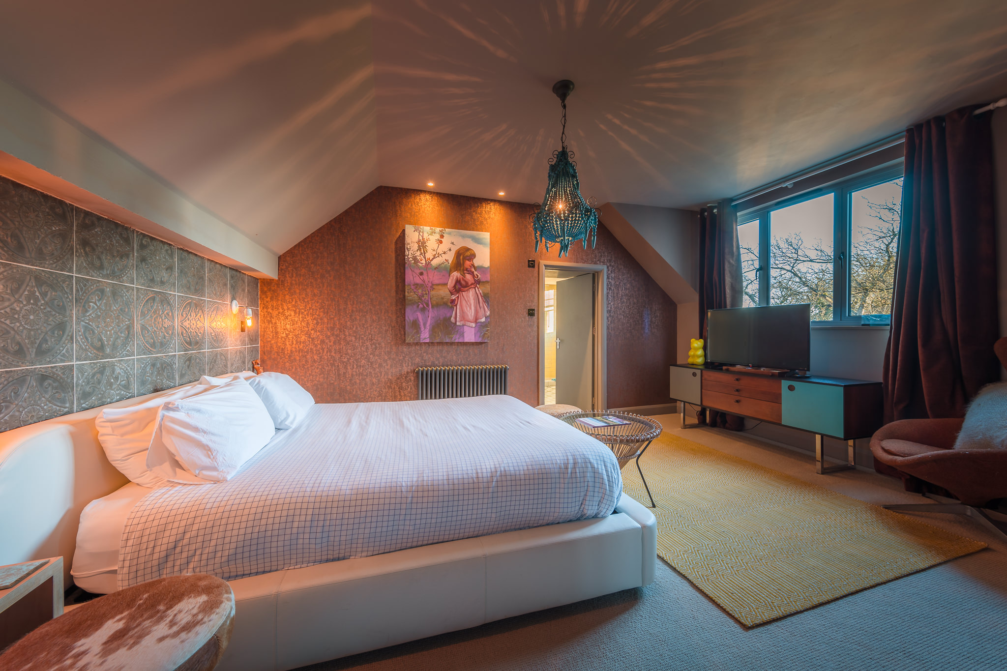 A cozy bedroom with a large white bed, bedside tables, patterned accent wall, chandelier, artwork above the bed, a TV on a console, large window with dark curtains, and warm ambient lighting. A cozy bedroom with a large white bed, bedside tables, patterned accent wall, chandelier, artwork above the bed, a TV on a console, large window with dark curtains, and warm ambient lighting.
