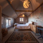 A cozy attic bedroom with a double bed, wooden furniture, patterned wallpaper, a rug with geometric designs, exposed beams, and large windows letting in natural light.