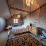 A cozy attic bedroom with colorful paisley wallpaper, exposed wooden beams, a double bed, wooden dresser, patterned rug, and a window letting in natural light.