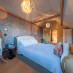 A cozy bedroom with a double bed, colorful paisley wallpaper, an ornate mirror, patterned pillows, and soft lighting from modern pendant lamps. Wooden beams and a small window add to the rooms warm, inviting feel.