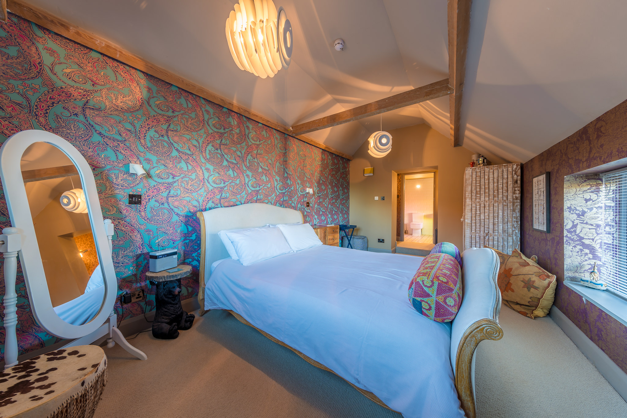 A cozy bedroom with a double bed, colorful paisley wallpaper, an ornate mirror, patterned pillows, and soft lighting from modern pendant lamps. Wooden beams and a small window add to the rooms warm, inviting feel. A cozy bedroom with a double bed, colorful paisley wallpaper, an ornate mirror, patterned pillows, and soft lighting from modern pendant lamps. Wooden beams and a small window add to the rooms warm, inviting feel.