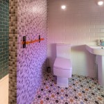 Modern bathroom with geometric patterned floor tiles, a white toilet, pedestal sink, and walls covered in white, green, and gray tiles. Two spotlights illuminate the sink and shelf above it.