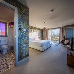 Bright modern bedroom with a white bed, floral accent wall, and open doors leading outside. An en-suite bathroom with patterned floor tiles and a toilet is visible to the left. Natural light fills the room.