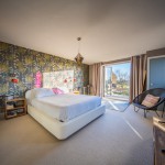 Bright bedroom with patterned wallpaper, a white bed, bedside tables, modern lighting, TV, chairs, and large glass doors opening to a balcony with a scenic outdoor view. Sunlight fills the room.