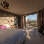 A cozy bedroom with a patterned accent wall, a large bed, a black chair, and double doors open to a balcony overlooking a sunny outdoor scene with trees and greenery. Warm light fills the room.