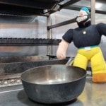 A plush toy figure dressed as a commis chef in yellow pants and a black shirt stands next to a metal frying pan on a stainless steel kitchen stove with grills and burners in the background.