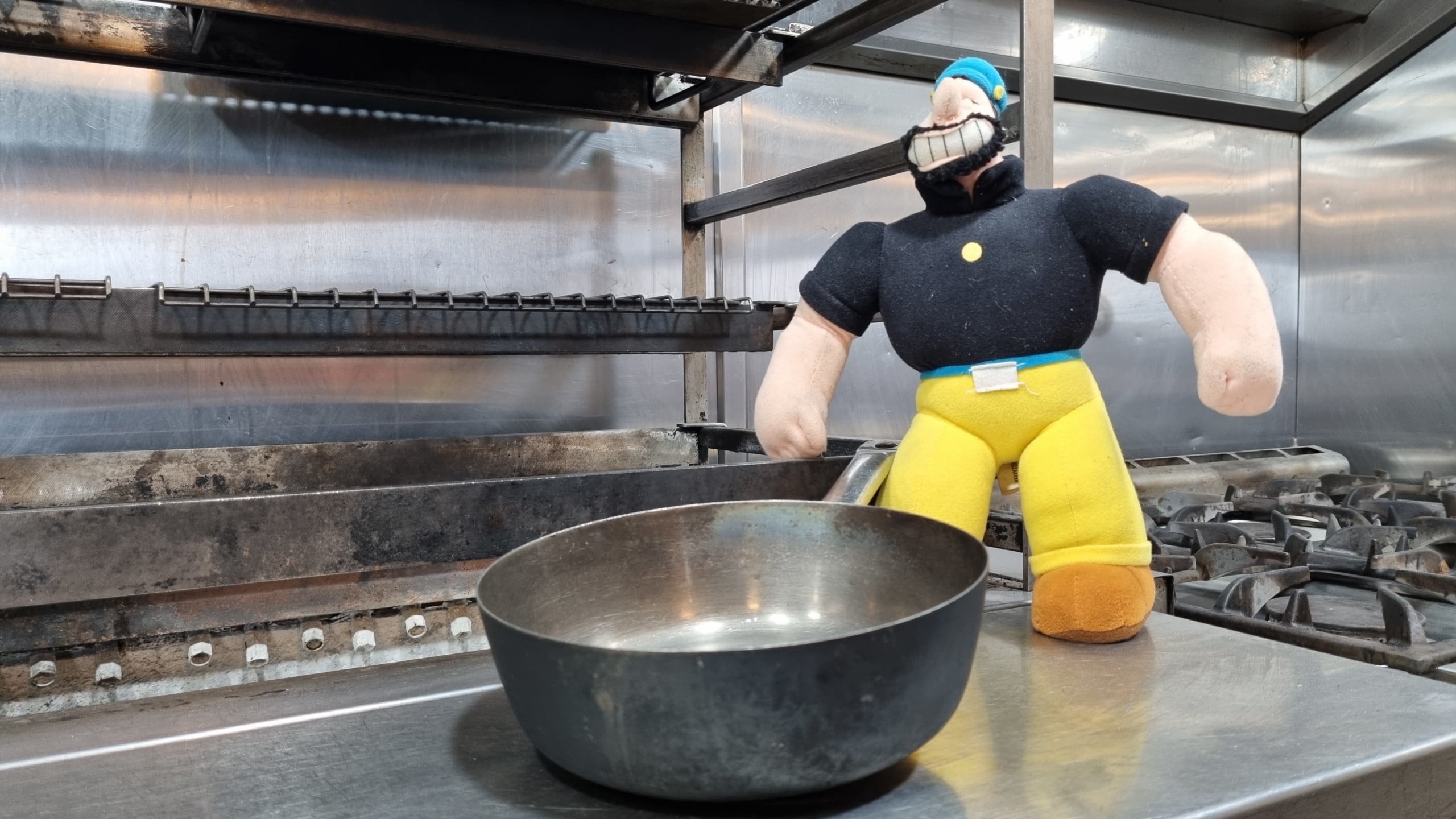 A plush toy figure dressed as a commis chef in yellow pants and a black shirt stands next to a metal frying pan on a stainless steel kitchen stove with grills and burners in the background.