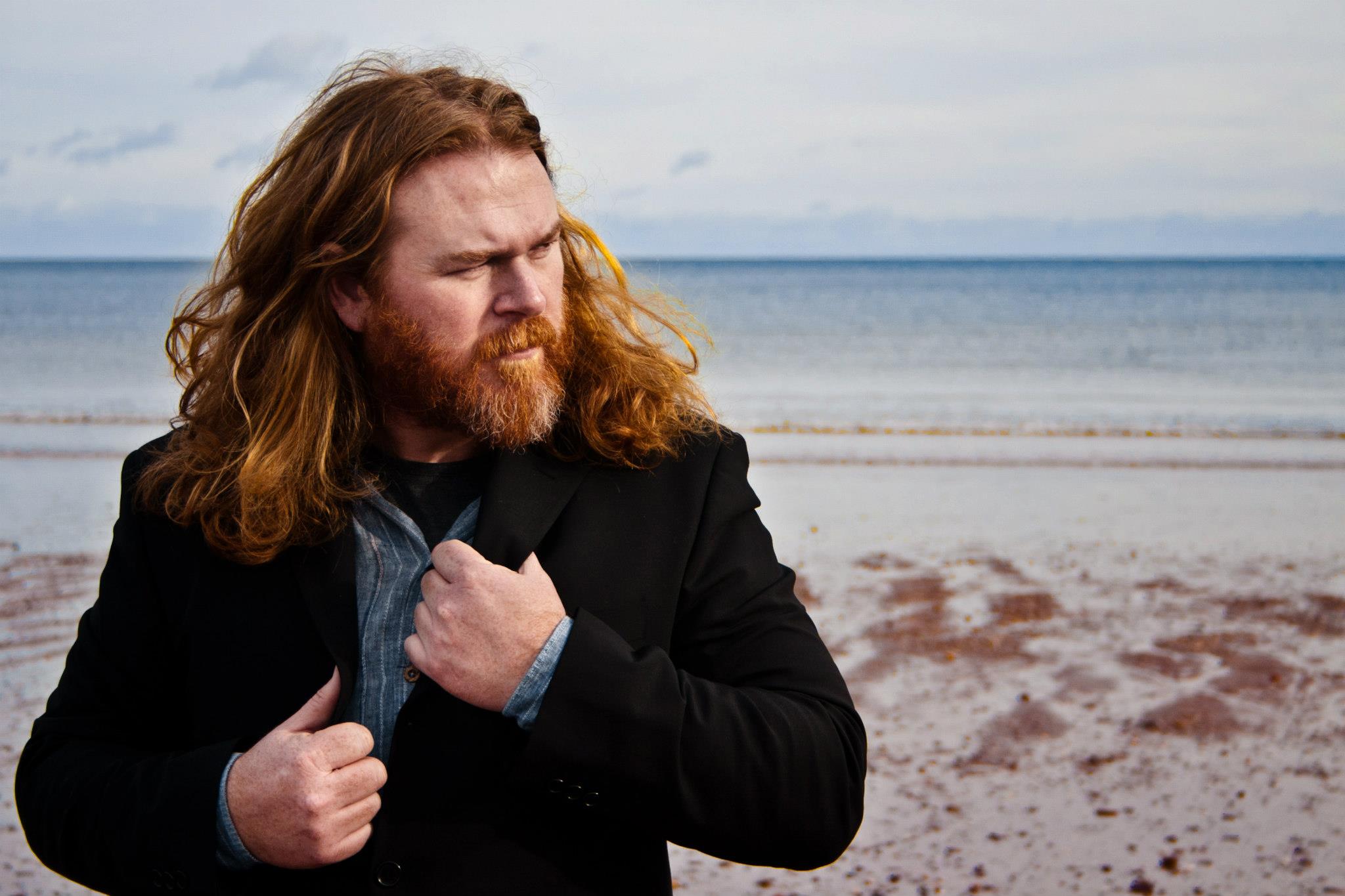 A man with long red hair and a beard, known as the Hairy Food Guy, stands on a sandy Sheppey beach near the water in a black jacket over a gray shirt, looking off to the side with a thoughtful expression as if pondering his next review.