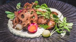 A gourmet dish featuring seared fish, crispy lotus root chips, microgreens, and pickled vegetables, artfully arranged on a radial dark plate with a milk stout-infused creamy herb puree and vibrant sauce.