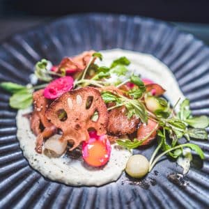 A plated gourmet dish featuring grilled meat, crispy lotus root chips, pickled vegetables, microgreens, and a creamy milk stout-infused white sauce on a dark, textured plate.