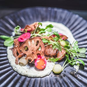 A gourmet dish featuring seared meat, lotus root chips, pickled vegetables, and greens on a swirl of creamy milk stout sauce, artfully arranged on a dark, textured plate.