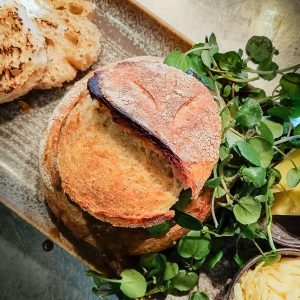 A close-up of crusty bread served on a plate with fresh green watercress, a slice of lemon, and a small bowl of grated cheese on the side. The golden, toasted bread pairs perfectly with a rich milk stout.