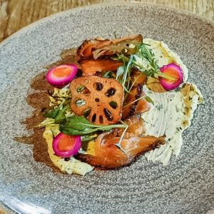 A gourmet dish featuring thinly sliced smoked meat, vibrant pickled quail eggs, fresh greens, and crispy lotus root chips on a swirl of creamy milk stout sauce, served on a textured gray plate.