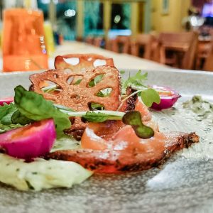 A close-up of a plated gourmet dish featuring smoked salmon, microgreens, a lotus root crisp, colorful pickled vegetables, and a creamy milk stout sauce, set on a gray plate in a warmly lit restaurant.