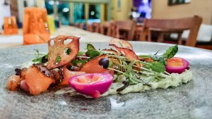 A gourmet dish featuring smoked salmon, microgreens, purple quail egg halves, and crispy lotus root chips, artfully arranged on a grey plate, perfectly paired with a rich milk stout in a refined restaurant setting.