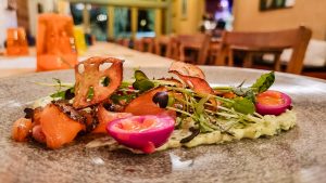 A close-up of a colorful gourmet dish featuring grilled salmon, microgreens, vibrant pink halved eggs, crispy lotus root chips, and creamy milk stout sauce on a gray plate in a warmly lit restaurant.