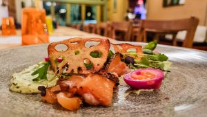 A plated gourmet dish featuring slices of cured fish, crispy lotus root chips, a halved pickled egg, microgreens, and a dollop of creamy herb sauce, set on a speckled plate in a cozy restaurant, perfectly paired with a rich milk stout.