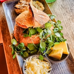 A rustic bread bowl filled with soup, topped with fresh greens, served with slices of crusty bread, a pile of grated cheese, and blocks of butter on a rectangular plate—perfectly paired with a rich milk stout for a cozy meal.