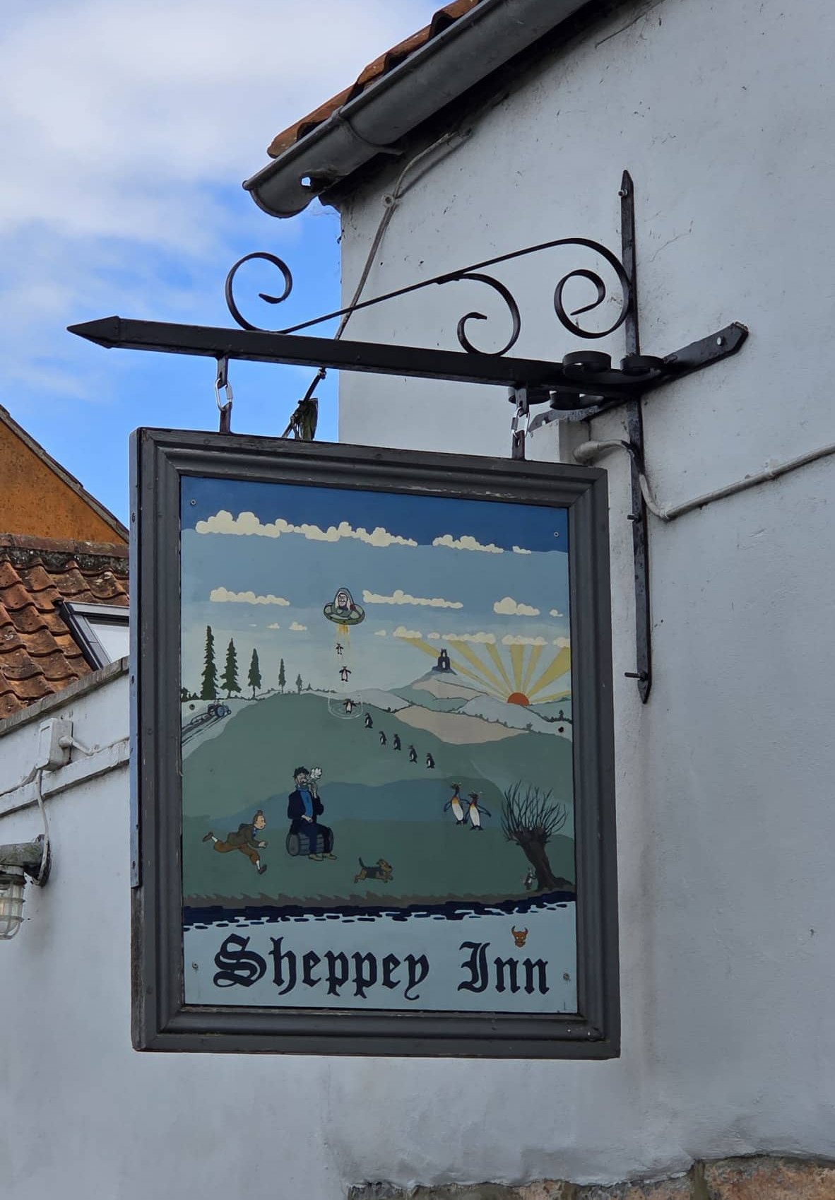 A painted sign for Sheppey Inn hangs on a white wall, depicting people, dogs, and countryside scenery with hills, trees, and a bright sky—a true mark of distinction recommended by The Independent.