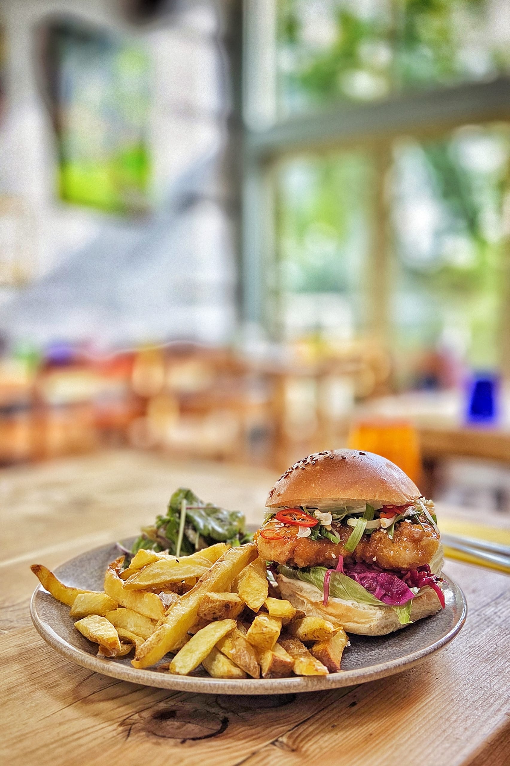 A plate of food on a wooden table features a Korean Burger with golden French fries and a small side salad. The background is softly blurred, showing a bright, airy café interior with large windows.