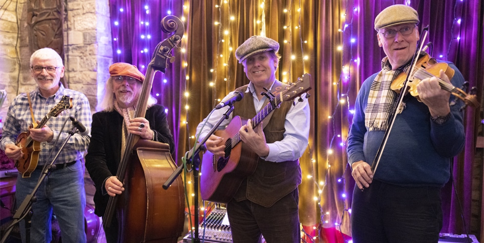 Four older men perform live music on stage in Boston, smiling as they play string instruments. They wear hats and casual clothing, with fairy lights and brown curtains, creating a warm, festive, tea party atmosphere.